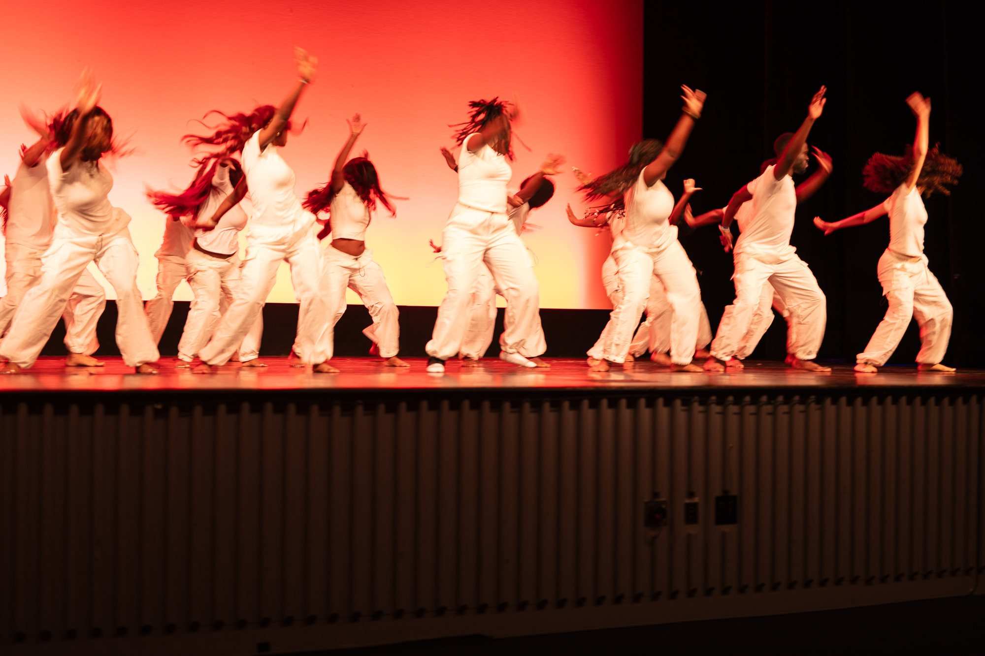 Dancers in white pose on a stage lit in orange and red