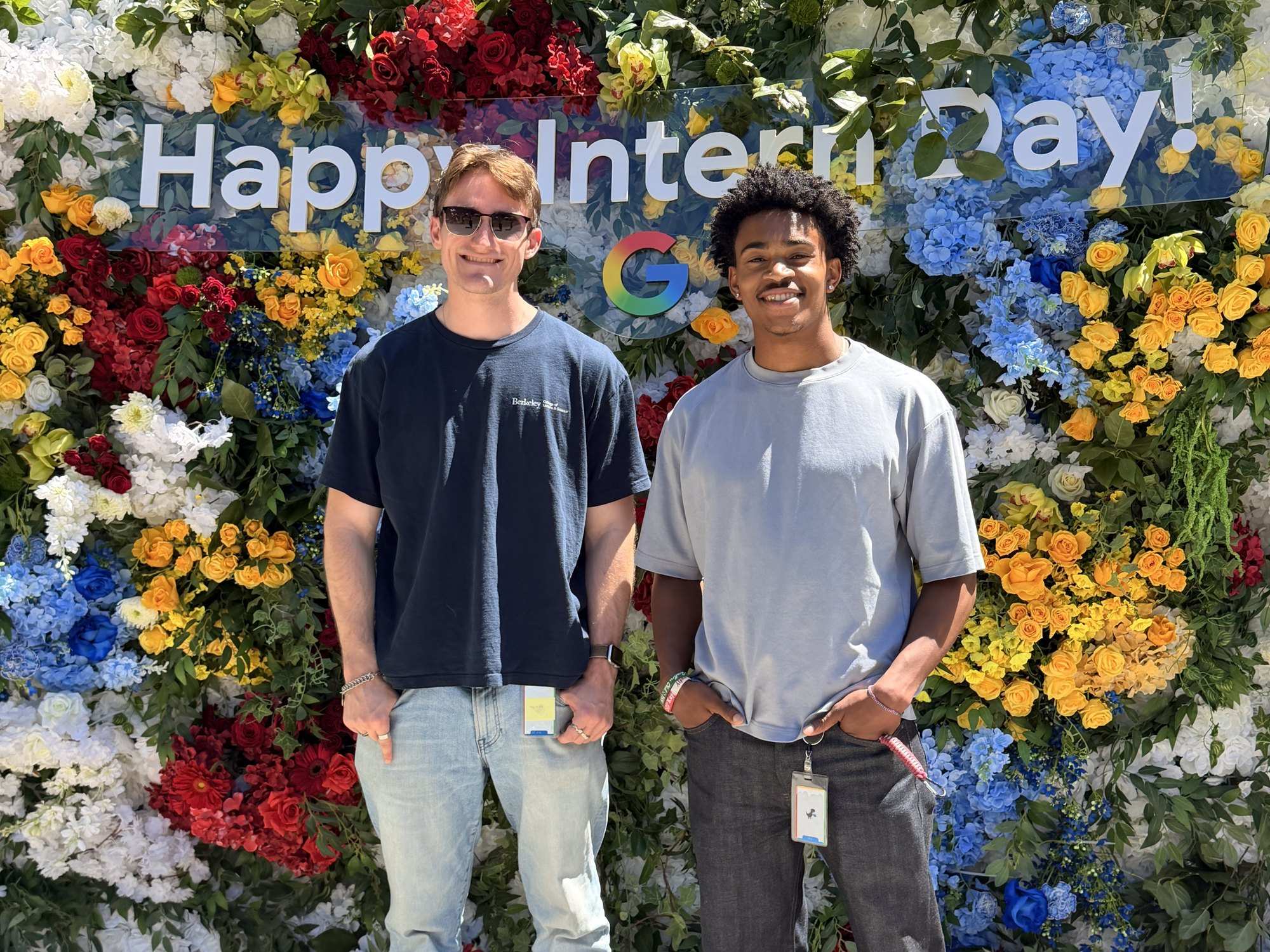 Two young men stand in front of a wall of flowers under a Happy Intern Day sign