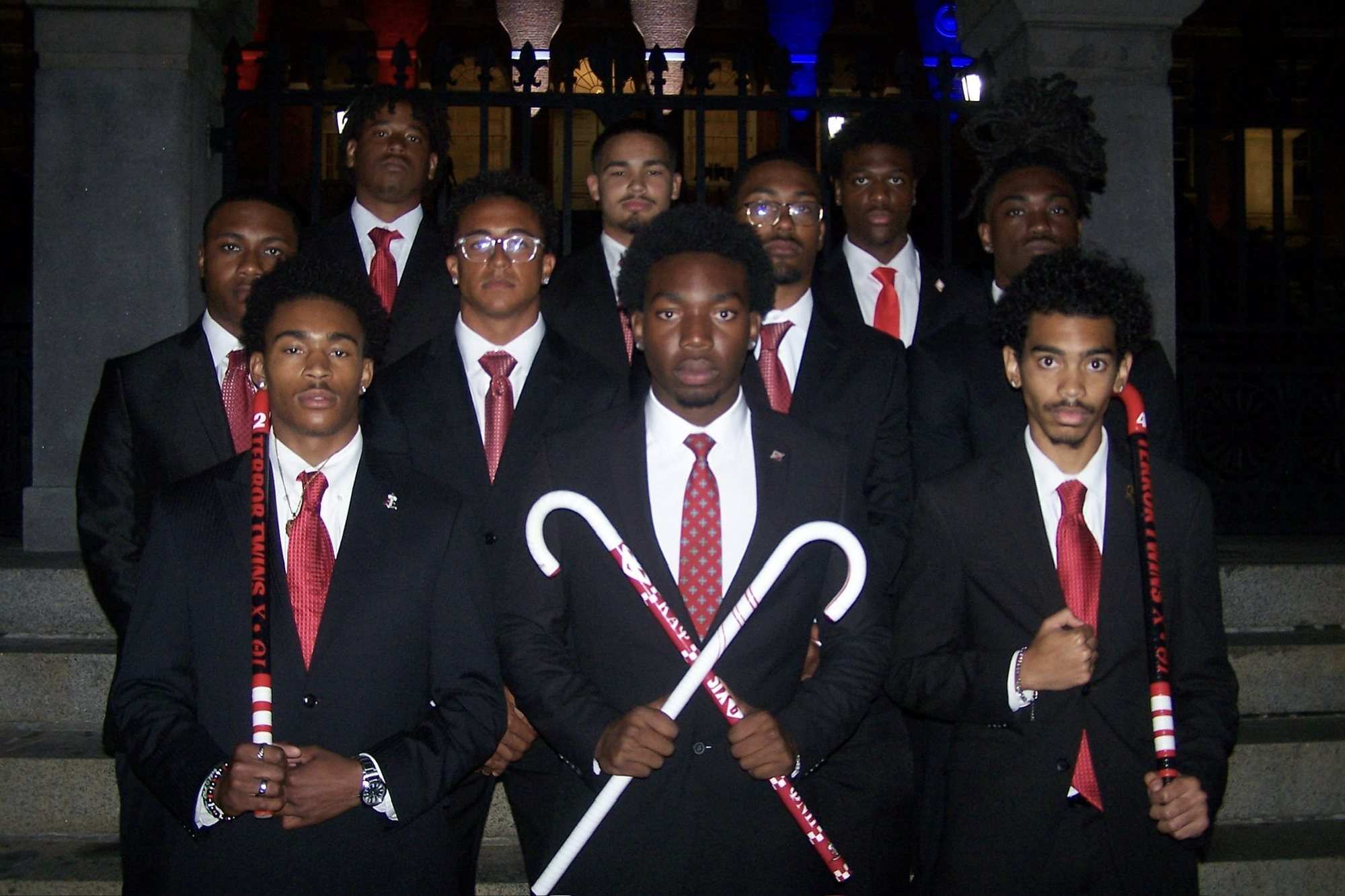 A group of men in dark suits and red ties stand together holding white canes at night in front of a stone building