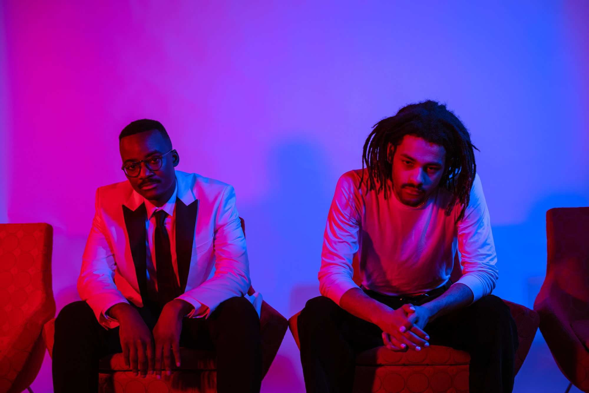 Two men seated in armchairs under magenta and blue light, one in a white suit jacket and tie, one in a white long-sleeved shirt