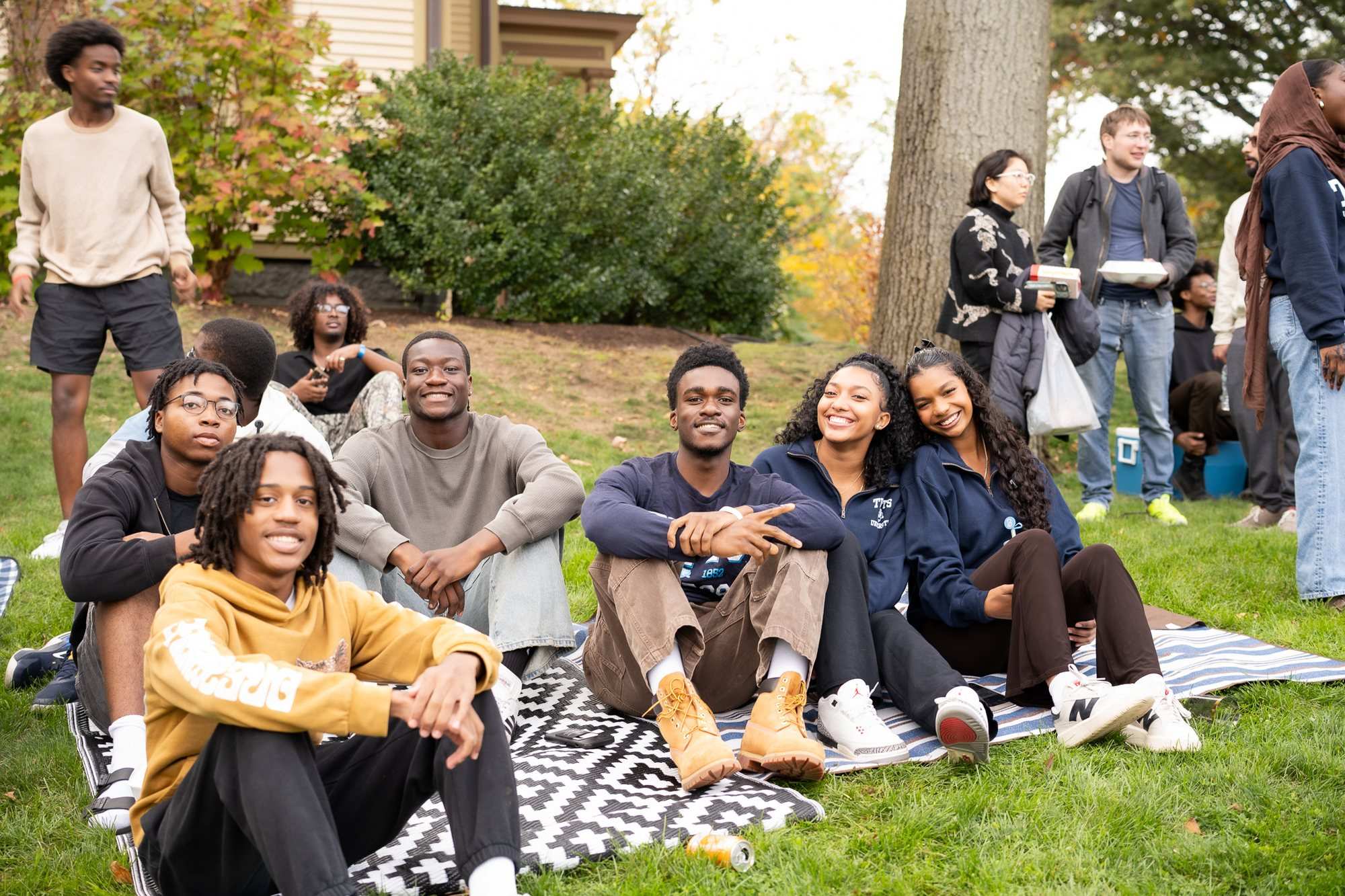 Students sit and stand together on a lawn among autumn trees, some on a patterned blanket, talking and smiling