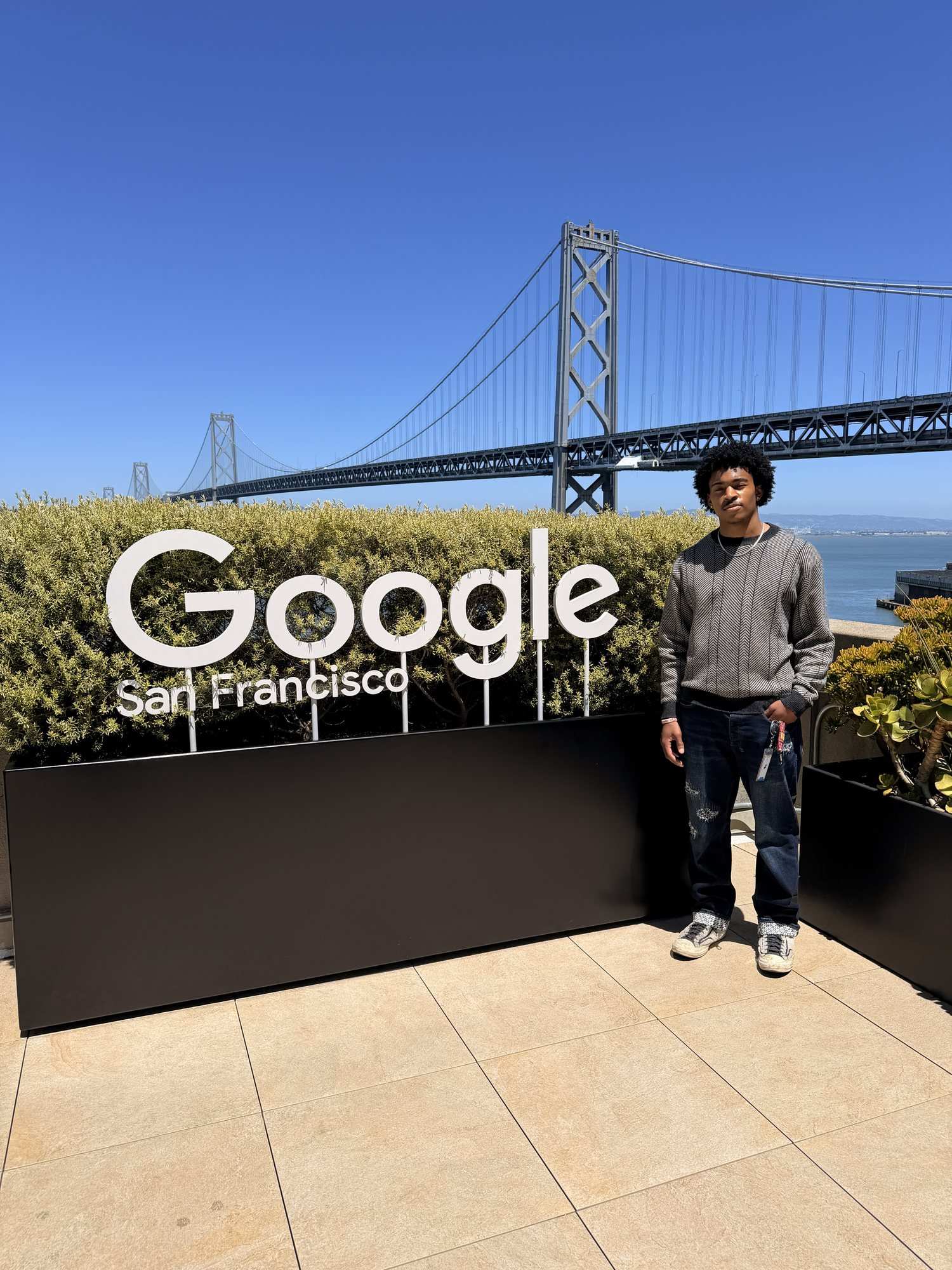 A man in a grey sweatshirt stands beside a Google San Francisco sign with a bridge and blue sky behind him