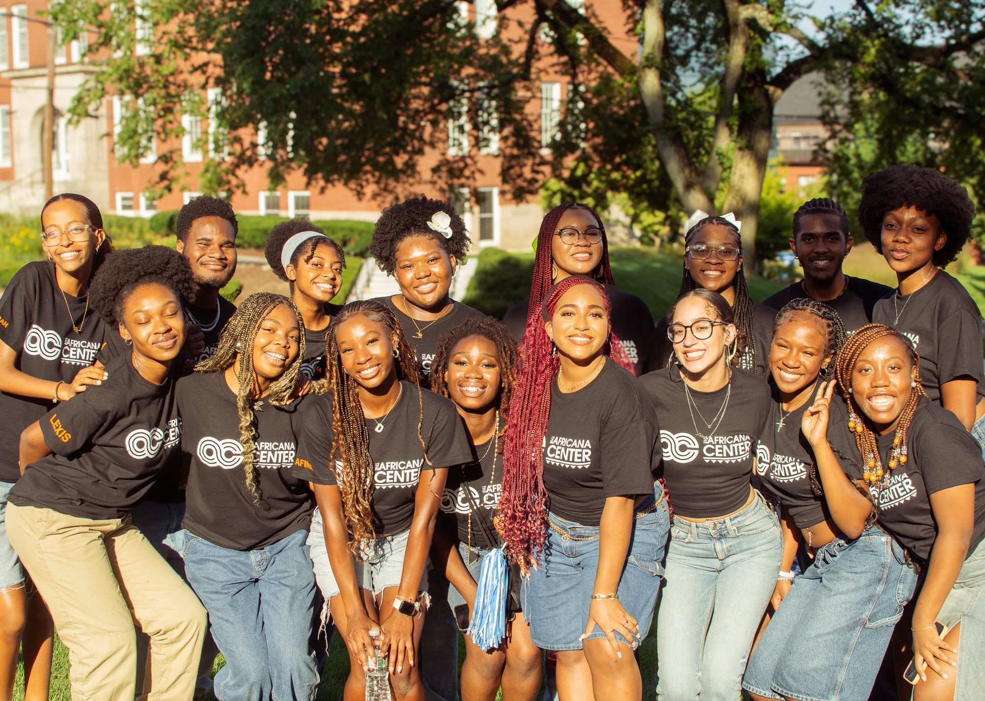 A large group of young people in matching black T-shirts pose close together outdoors in front of brick buildings
