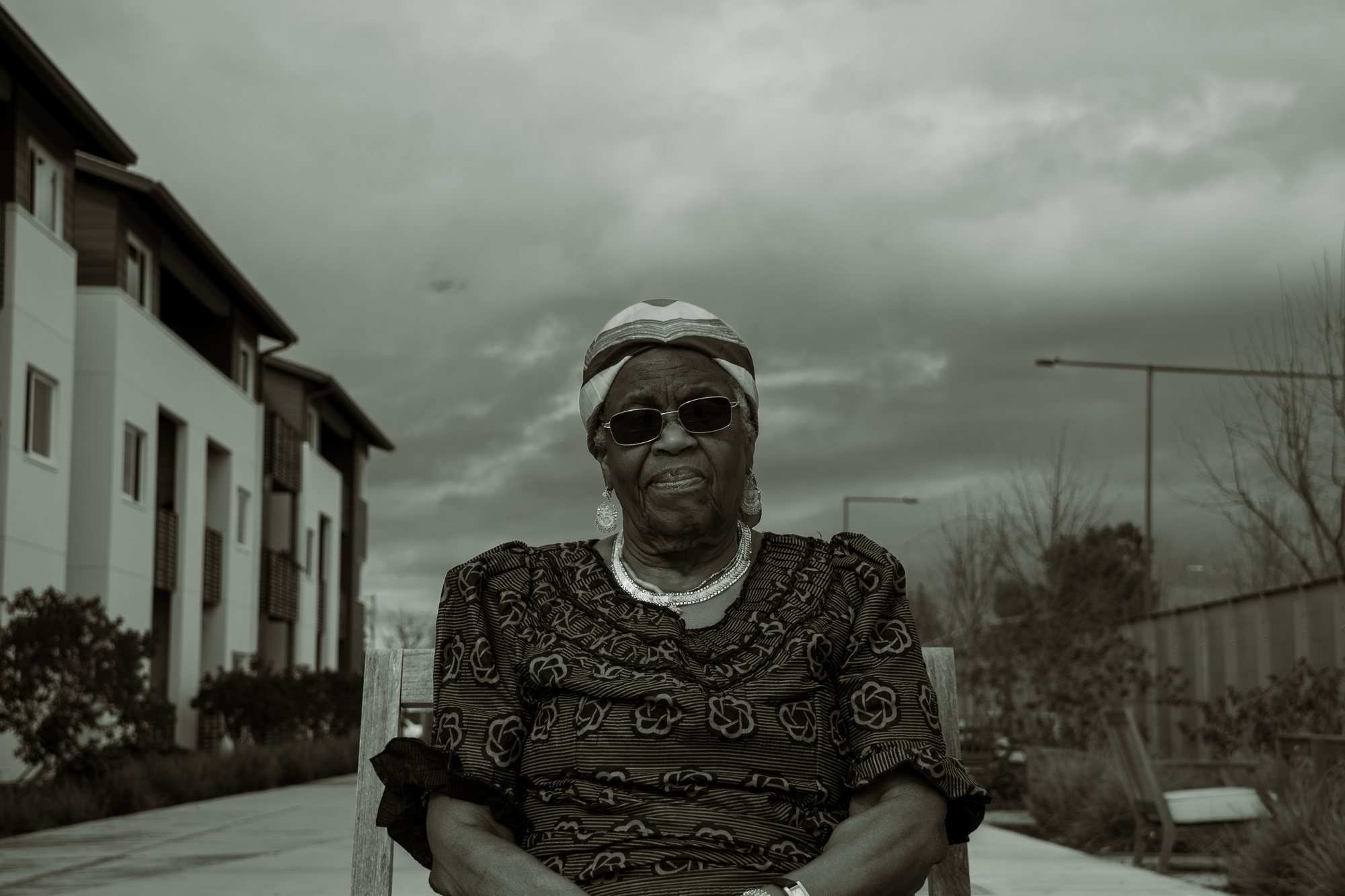An older woman in sunglasses, a patterned dress and a head wrap, seated outdoors under a cloudy sky