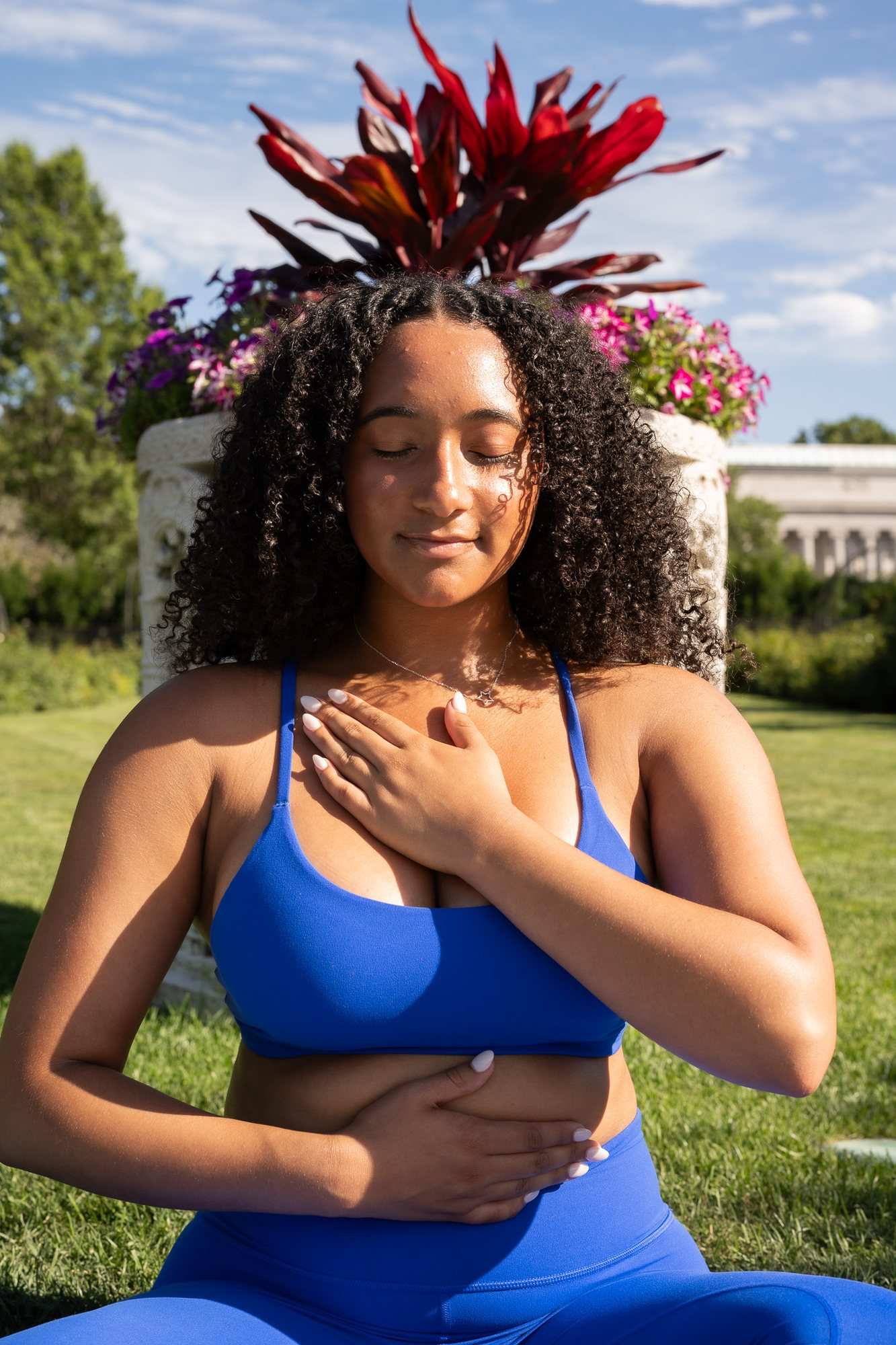 A woman in a blue sports set sits on grass with her eyes closed and her hands on her chest and stomach