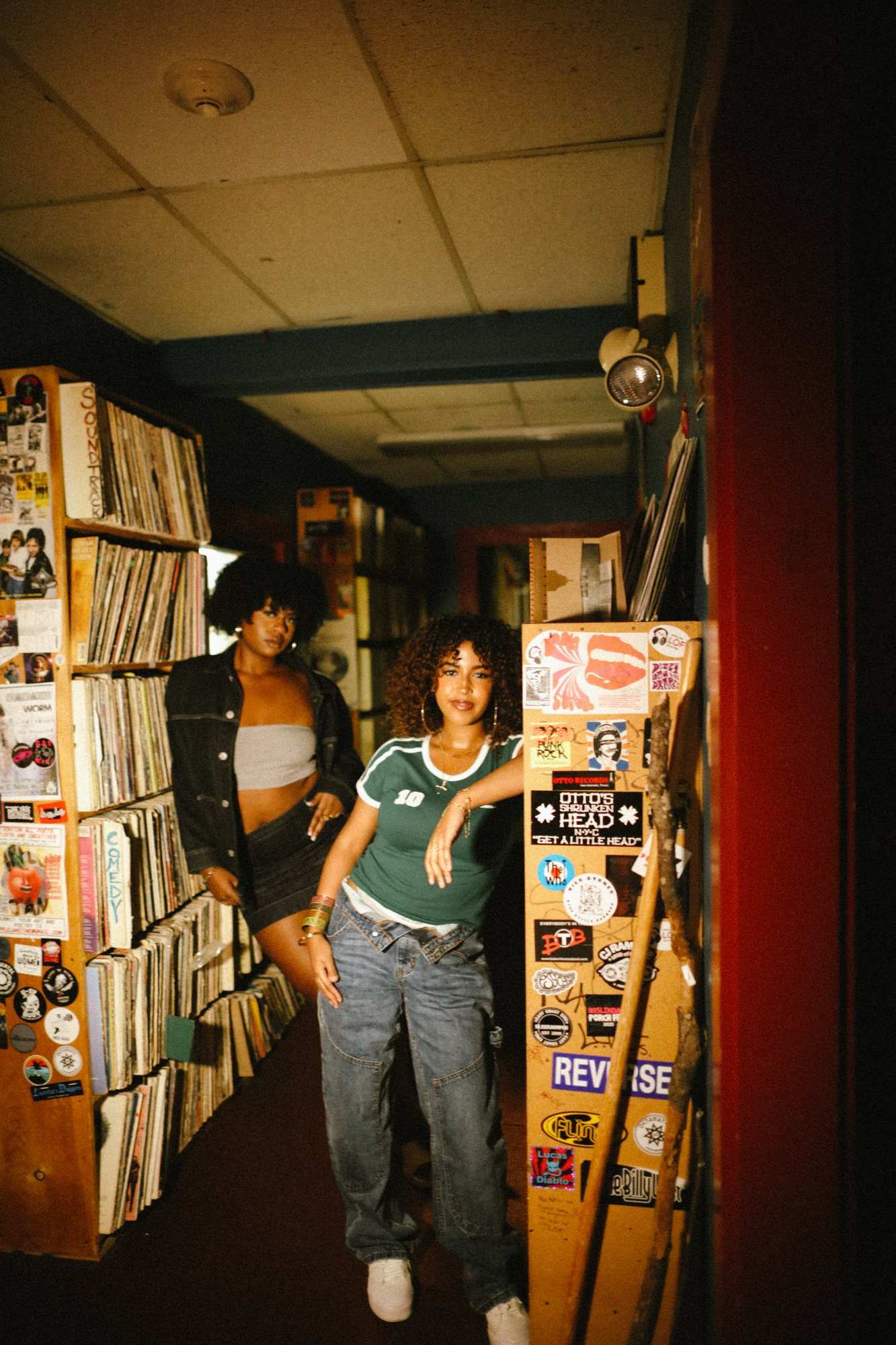 Two young women posing in the narrow aisle of a record store, between shelves of vinyl and a sticker-covered pillar