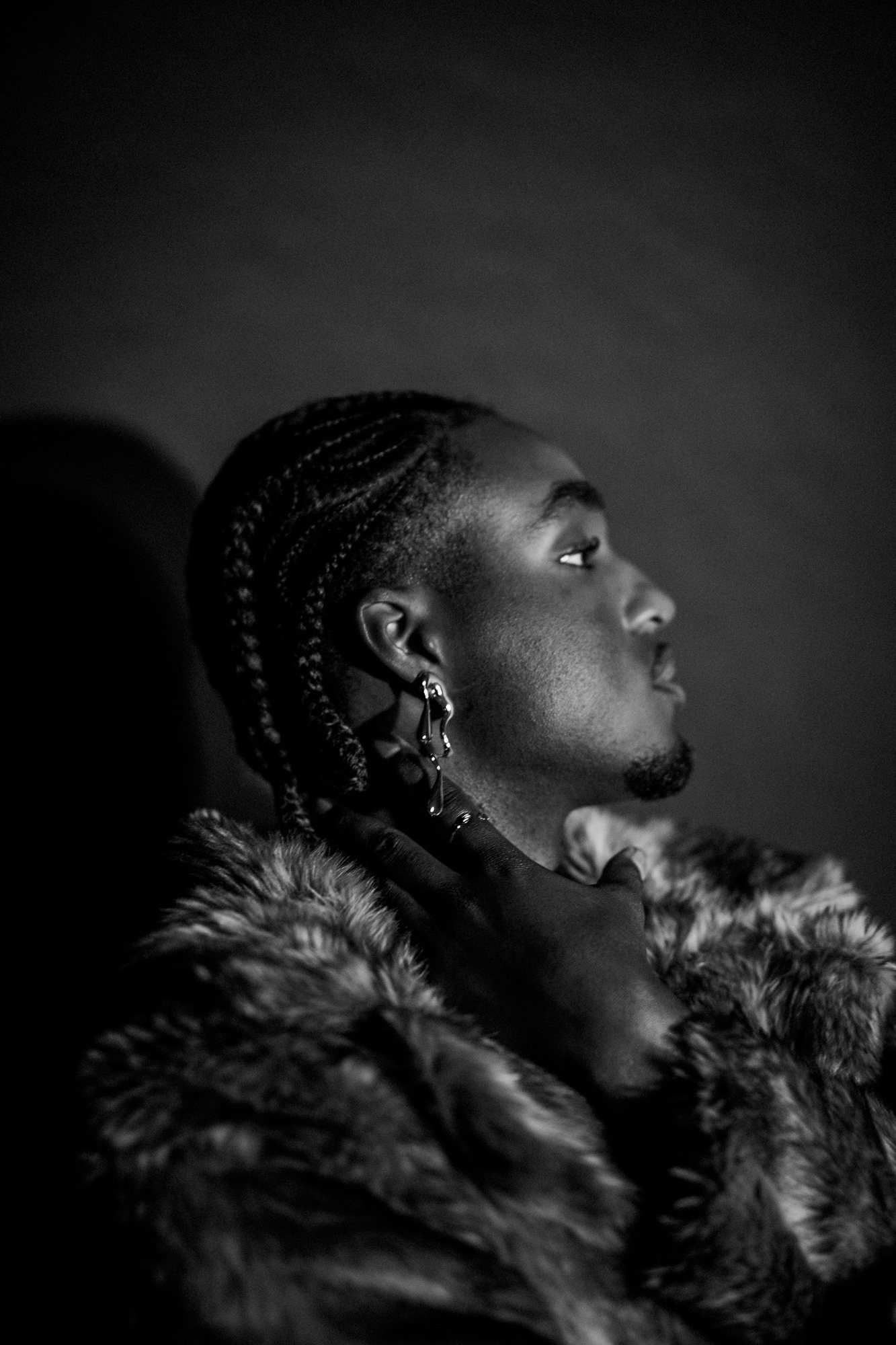 Black and white portrait of a man in profile with braided hair, an earring and a fur coat